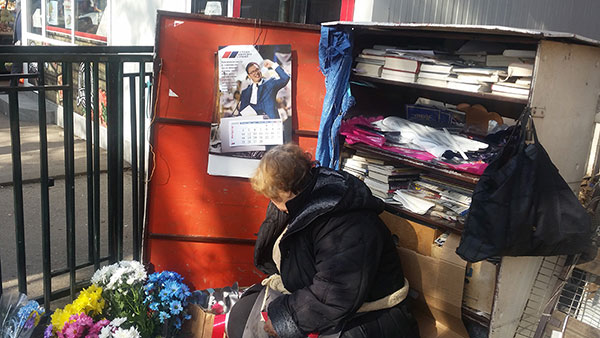 žena na pijaci prodaje cveće, a iza nje je kalendar sa likom Vučića